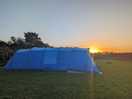 Large blue family tent in camping field with sun setting behind it.
