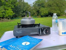 Camping table with small gas stove, kettle and book and views across a quiet camp site
