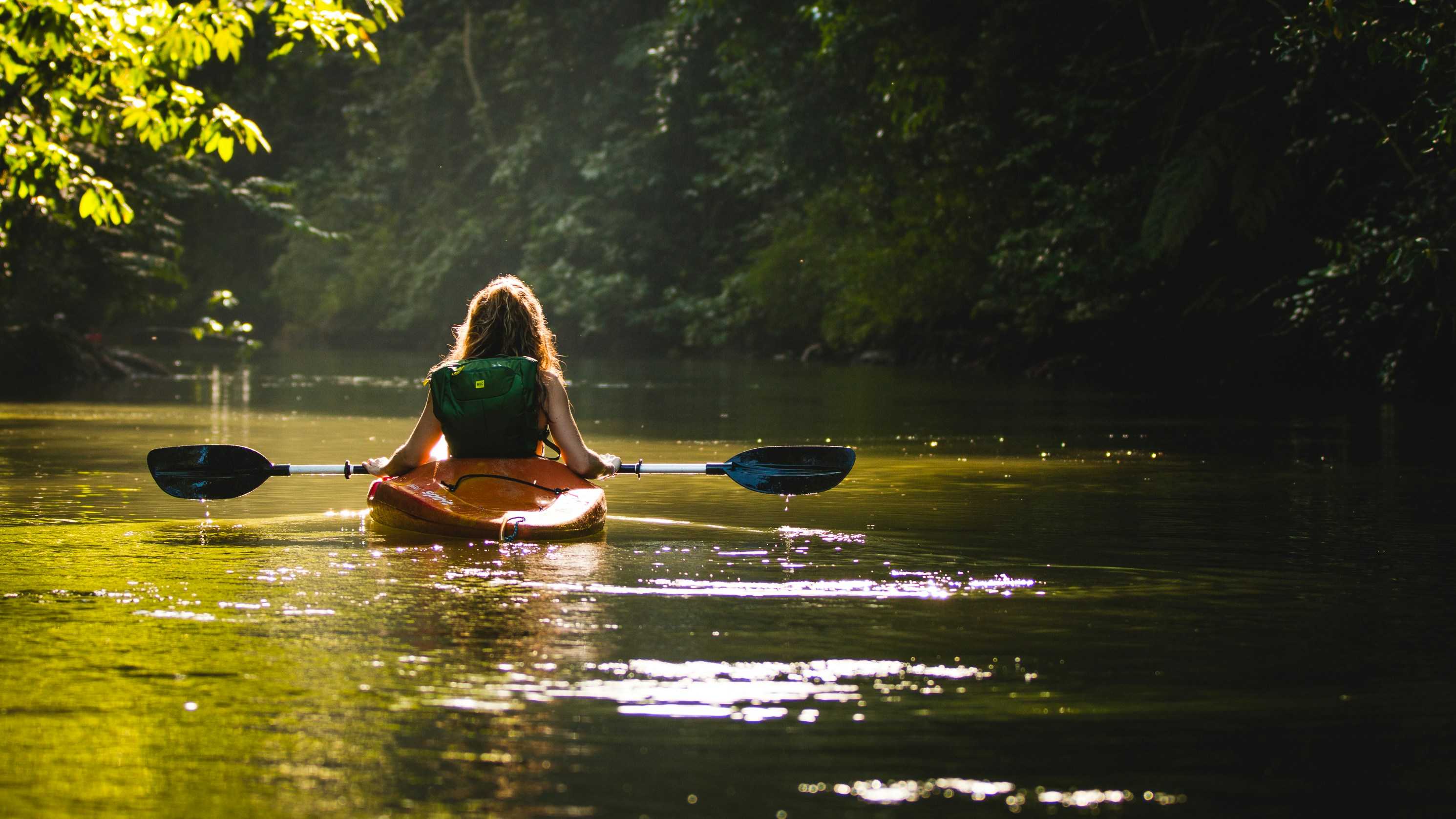Image shows a woman with her back to the camera kayaking down a river with green trees at either side