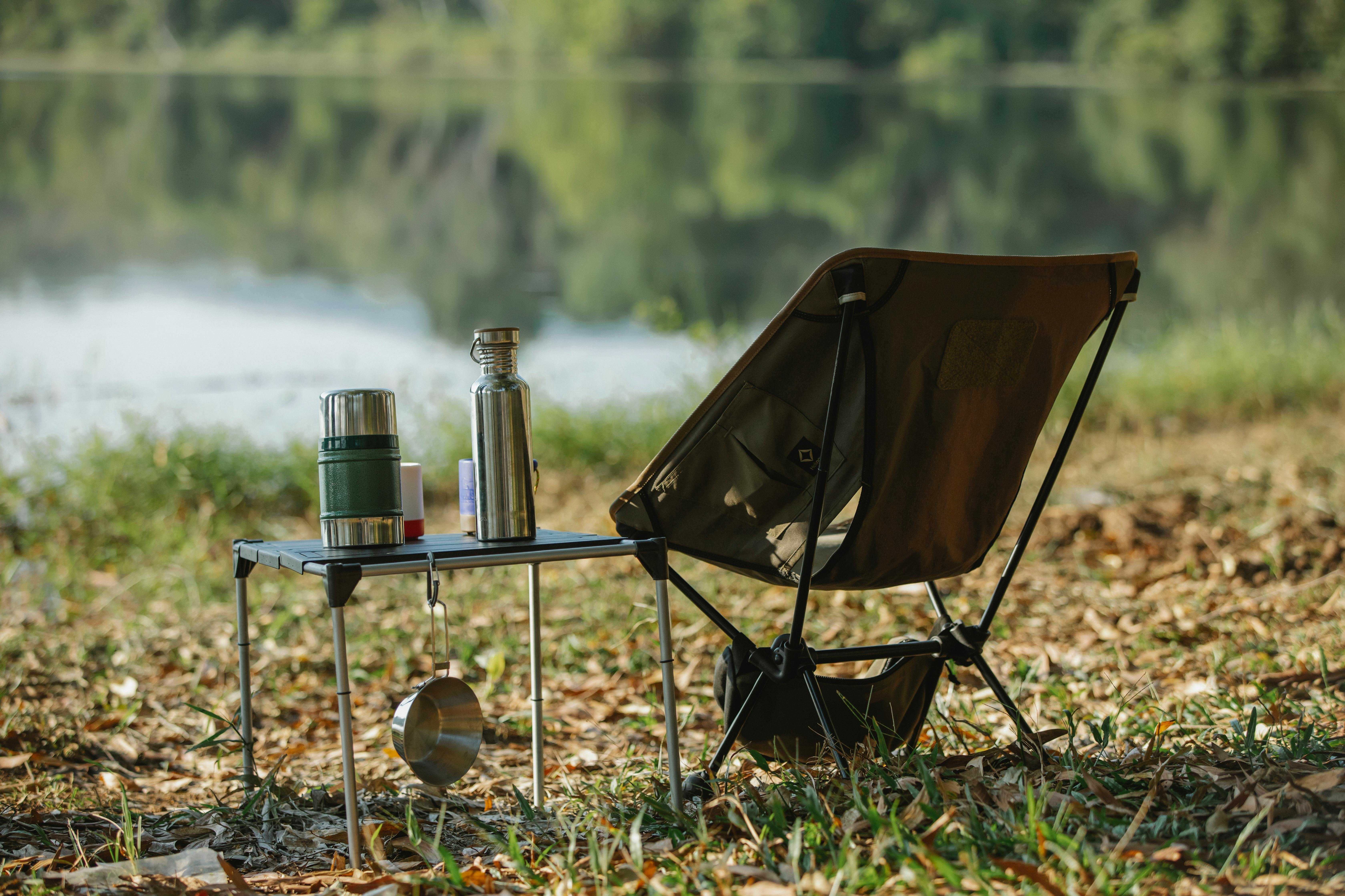 An empty camping chair looks out over a river, with a flask of coffee on a table beside